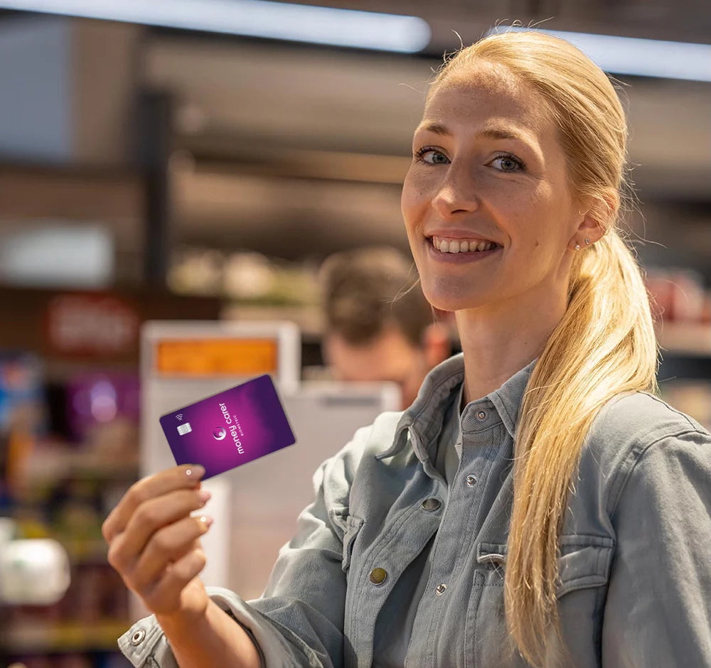 a woman is in a grocery store. she is in focus. she is looking forwards and smiling. in her hand she is holding a purple and pink gradient card with the money carer logo on.