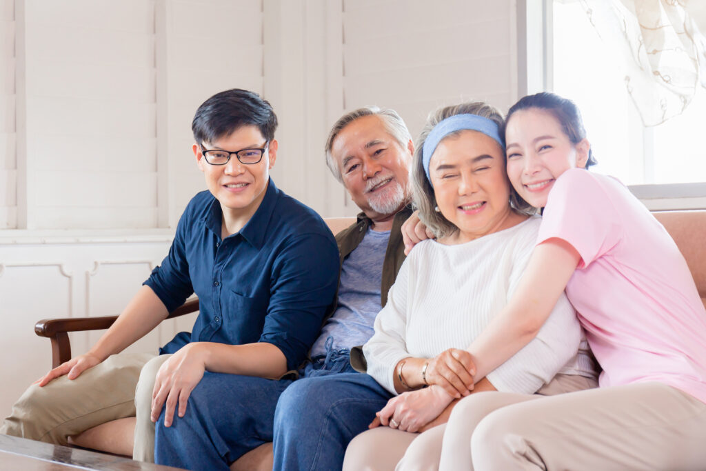 a family are sitting on a sofa together indoors. the parents are sat in the middle, the son is on the left, the daughter is on the right. they are all hugging and smiling. the parents are elderly.