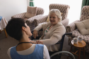 a nurse is talking to an elderly woman in a wheelchair. she is holding the woman's wrist, as if checking her pulse. there are flowery armchairs behind them, and a table next to the elderly woman with two cups of coffee.