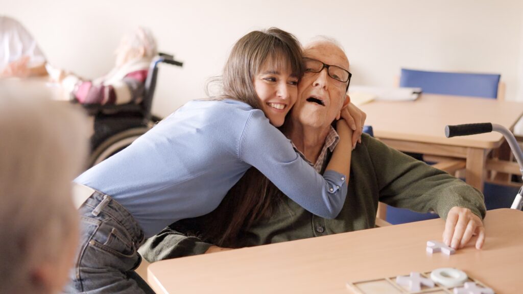 a young woman is kneeling by the side of her elderly grandfather and smiling. they appear to be in a care home. the elderly man is sat at a table playing a game of noughts and crosses.