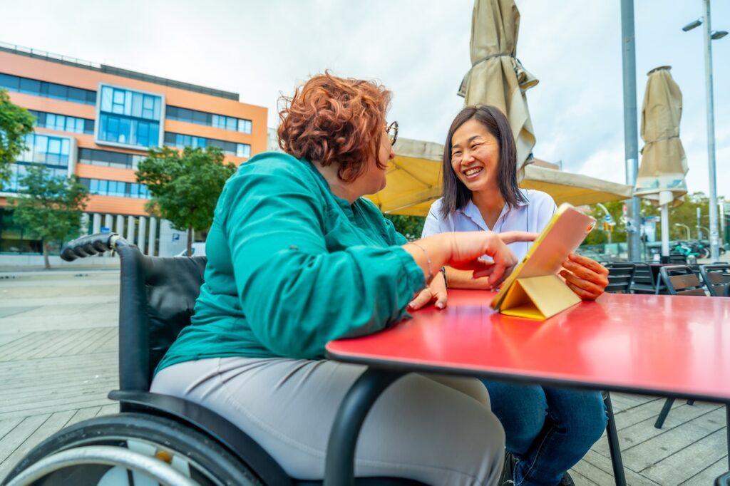 two women are sat discussing at a table outside. the woman on the left is in a wheelchair. there is a digital tablet propped up on the table, that the woman in the wheelchair is pointing at. both women are looking at each other and smiling.